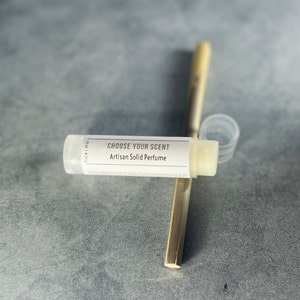 May include: A white tube of solid perfume with the label "Choose Your Scent Artisan Solid Perfume". The tube is 1/2 oz and is next to a wooden stick.