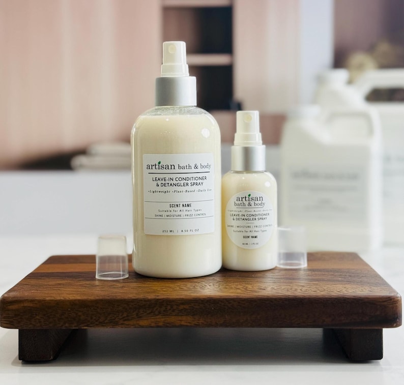May include: Two bottles of Artisan Bath & Body leave-in conditioner and detangler spray on a dark wooden tray. The bottles are white with silver spray nozzles. The text on the bottles reads "LEAVE-IN CONDITIONER & DETANGLER SPRAY".