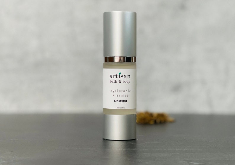 May include: A silver bottle with a white label that reads "artisan bath & body hyaluronic + arnica LIP SERUM 1 fl oz / 30 ml".