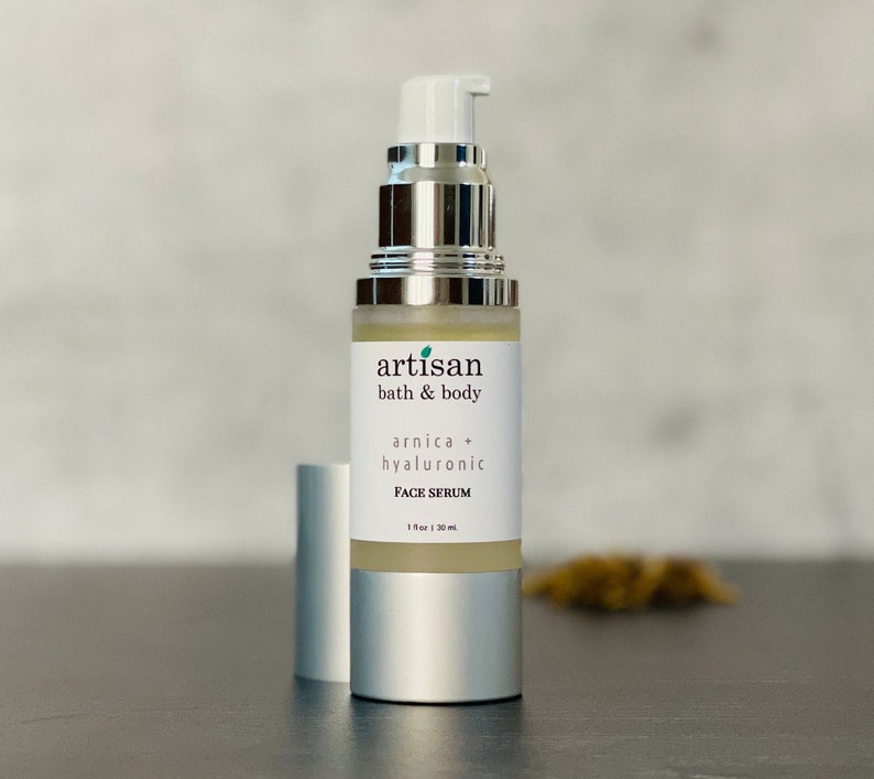 Op de afbeelding: Een zilveren fles met een heldere vloeistof erin. De fles is gelabeld met "artisan bath & body arnica + hyaluronic FACE SERUM 1 fl oz | 30 ml".