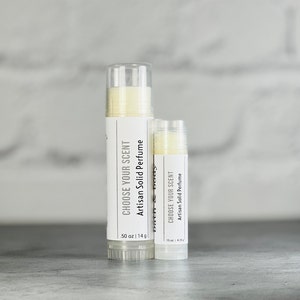 May include: Two white tubes of solid perfume with the text "Choose Your Scent Artisan Solid Perfume" and the size in ounces and grams. The larger tube is 50 oz | 14 g and the smaller tube is 15 oz | 4.25 g.