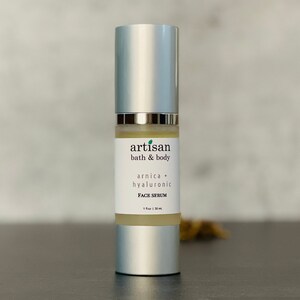 Op de afbeelding: Een zilveren fles met een doorzichtig etiket waarop staat "artisan bath & body arnica + hyaluronic FACE SERUM 1 fl oz | 30 ml".