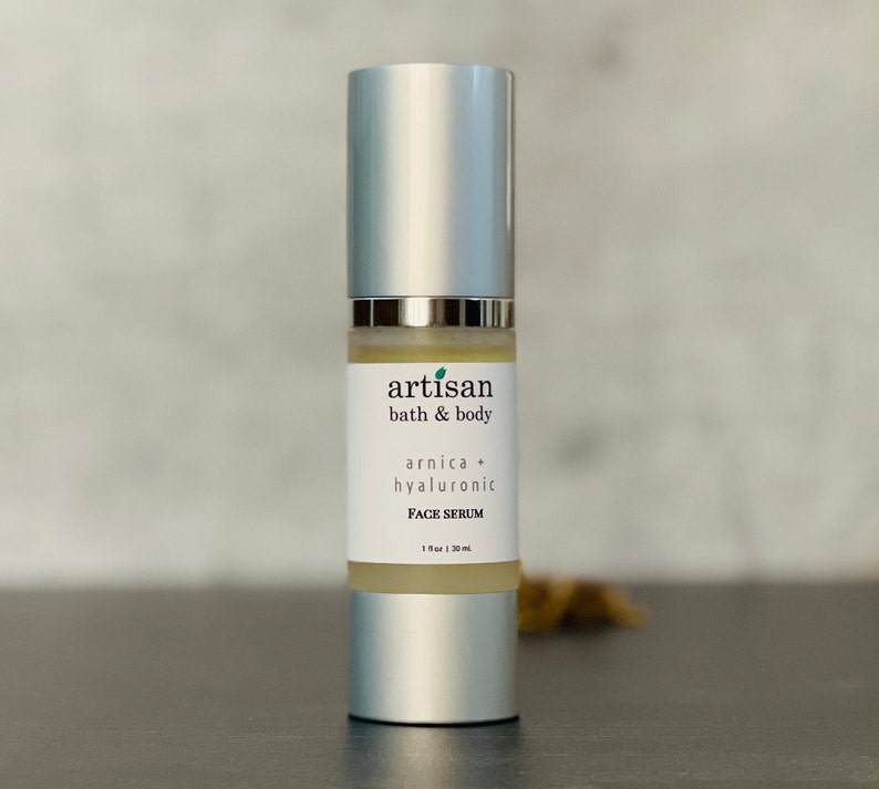 Op de afbeelding: Een zilveren fles met een doorzichtig etiket waarop staat "artisan bath & body arnica + hyaluronic FACE SERUM 1 fl oz | 30 ml".