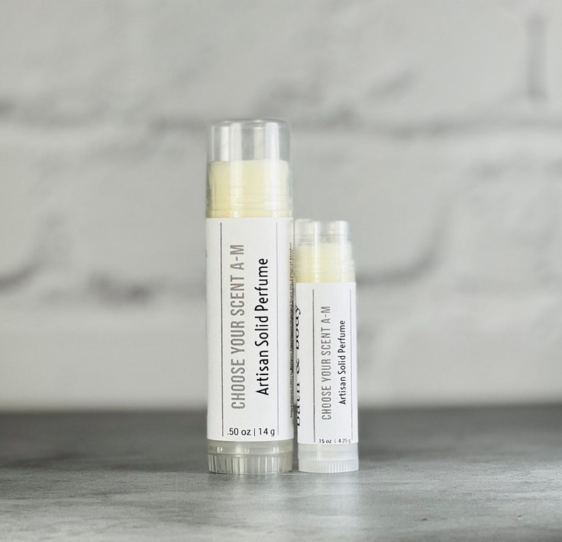 May include: Two white tubes of solid perfume, one larger than the other. The larger tube is labelled "Choose Your Scent A-M Artisan Solid Perfume .50 oz | 14 g". The smaller tube is labelled "Choose Your Scent A-M Artisan Solid Perfume .15 oz | 4.25 g".