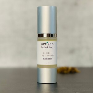 Op de afbeelding: Een zilveren flesje Arnica + Hyaluronzuur gezichts serum van Artisan Bath & Body. De fles is gelabeld met de merknaam en productbeschrijving. De fles is 1 fl oz / 30 ml.