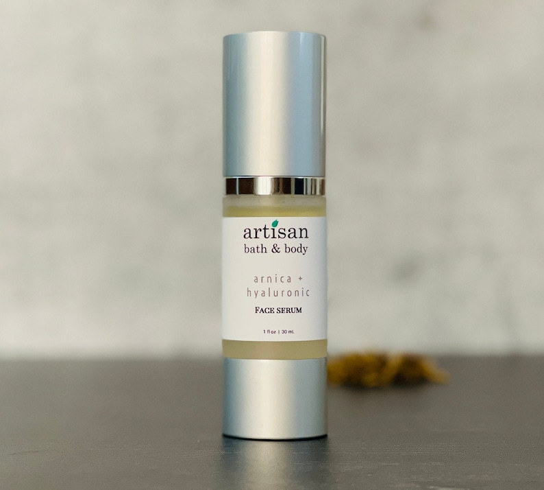 Op de afbeelding: Een zilveren flesje Arnica + Hyaluronzuur gezichts serum van Artisan Bath & Body. De fles is gelabeld met de merknaam en productbeschrijving. De fles is 1 fl oz / 30 ml.