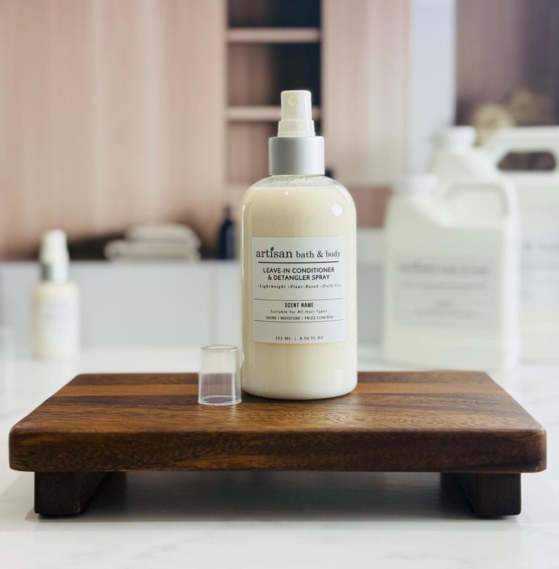 May include: A bottle of Artisan bath & body leave-in conditioner & detangler spray with a silver cap, sitting on a small wooden tray. The label indicates the product is lightweight, plant-based, and suitable for all hair types. The bottle is 8.5 fl oz.