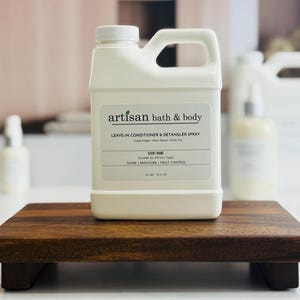 May include: A white plastic bottle of Artisan Bath & Body leave-in conditioner and detangler spray, with a handle and a white label. The label includes the product name, scent information, and volume of 16 fl oz. The bottle sits on a small, dark wooden tray.