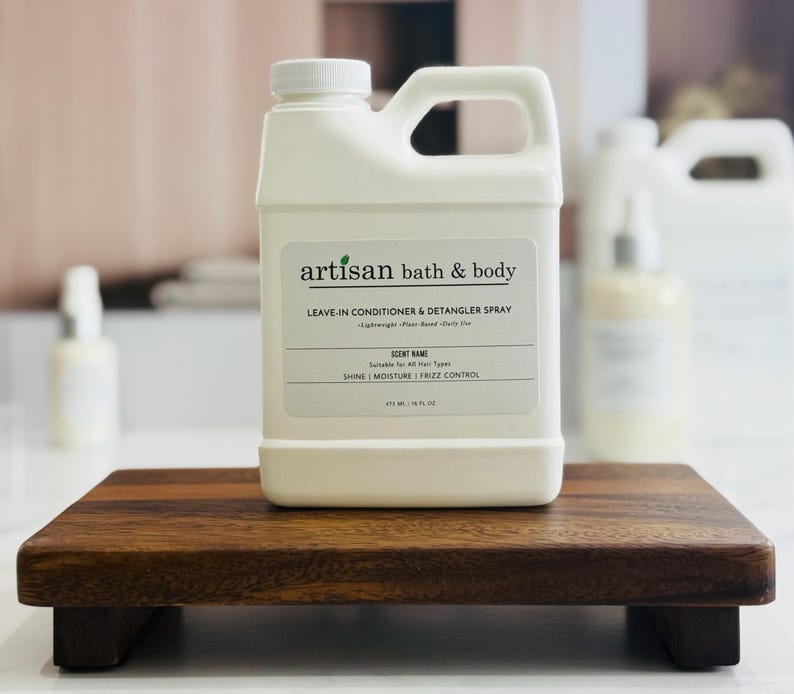 May include: A white plastic bottle of Artisan Bath & Body leave-in conditioner and detangler spray, with a handle and a white label. The label includes the product name, scent information, and volume of 16 fl oz. The bottle sits on a small, dark wooden tray.