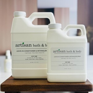 May include: Two white plastic bottles of Artisan Bath & Body leave-in conditioner and detangler, one large and one small, with black text labels. The labels include the product name, scent name, and details about the product. The bottles are on a dark wooden tray.