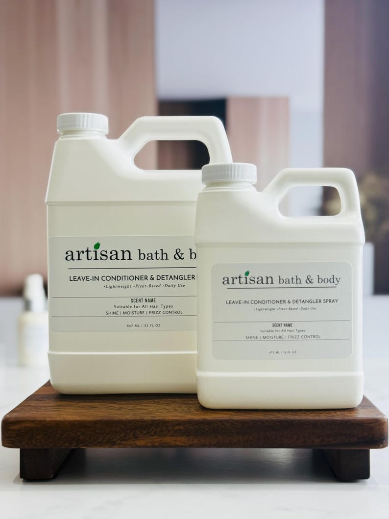 May include: Two white plastic bottles of Artisan Bath & Body leave-in conditioner and detangler, one large and one small, with black text labels. The labels include the product name, scent name, and details about the product. The bottles are on a dark wooden tray.