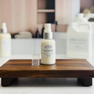 May include: A bottle of Artisan Bath & Body leave-in conditioner and detangler spray with a silver cap and a clear plastic cap, sitting on a small wooden tray. The label includes the words "Shine / Moisture / Frizz Control".