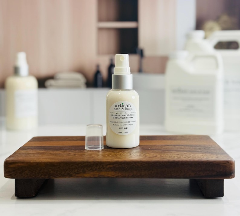 May include: A bottle of Artisan Bath & Body leave-in conditioner and detangler spray with a silver cap and a clear plastic cap, sitting on a small wooden tray. The label includes the words "Shine / Moisture / Frizz Control".