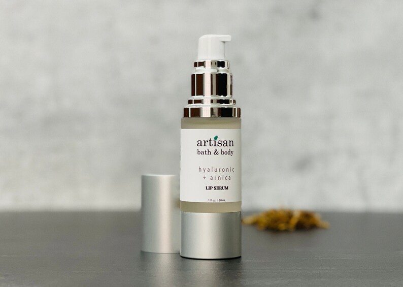 May include: A silver bottle with a clear liquid inside. The bottle is labeled "artisan bath & body hyaluronic + arnica lip serum 1 fl oz (30 ml)".