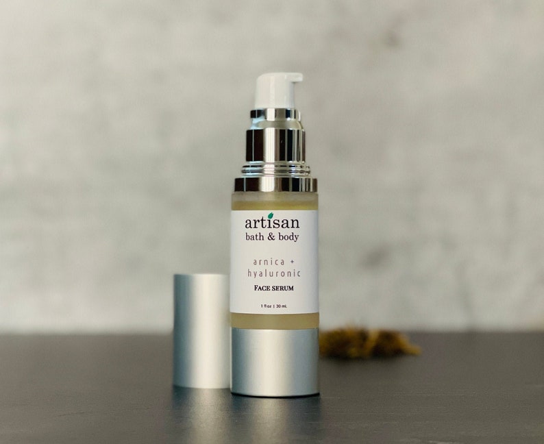 Op de afbeelding: Een zilveren fles met een pompdispenser die een heldere vloeistof bevat. De fles is gelabeld met "artisan bath & body arnica + hyaluronic FACE SERUM 1 fl oz / 30 ml".