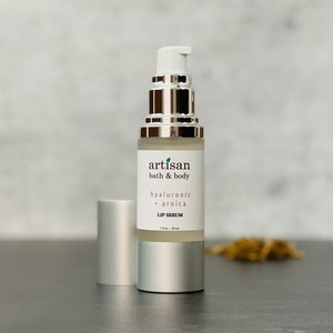May include: A silver bottle with a clear liquid inside. The bottle is labeled "artisan bath & body hyaluronic + arnica lip serum 1 fl oz | 30 ml".