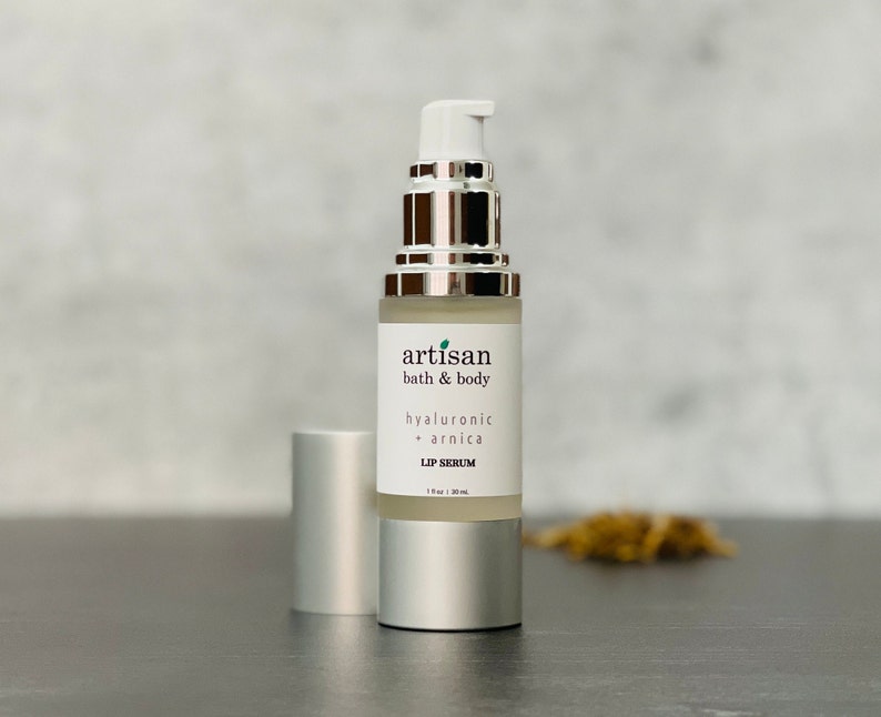 May include: A silver bottle with a clear liquid inside. The bottle is labeled "artisan bath & body hyaluronic + arnica lip serum 1 fl oz | 30 ml".