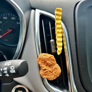 May include: A close-up of a car's interior with a brown chicken nugget and a yellow, wavy, air freshener hanging from the air vent. The car's dashboard has a speedometer, a rear window defroster button, and an engine start/stop button.