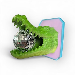 Pode incluir: Uma escultura de jacar&eacute; verde segurando uma bola de discoteca prateada. O jacar&eacute; &eacute; montado em uma placa retangular rosa e azul. A escultura tem uma apar&ecirc;ncia texturizada e escamosa.