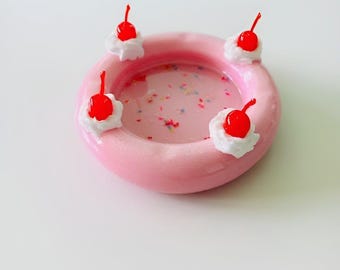 May include: A pink inflatable pool ring with white frosting and red cherries on top. The pool is filled with pink water and sprinkles.