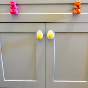 May include: A set of cabinet hardware featuring a pink bear, an orange bear, and two white egg-shaped knobs with yellow accents.