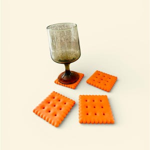 Puede incluir: Una copa de vino con base marrón y cuenco transparente y texturizado descansa sobre un posavasos cuadrado naranja. Tres posavasos cuadrados naranjas adicionales con diseño de galleta se dispersan alrededor de la copa.
