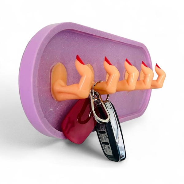 May include: A unique wall-mounted key holder featuring four sculpted hands with red painted fingernails. The hands extend from a lavender-colored rectangular base, holding keys. A creative and functional home decor item.
