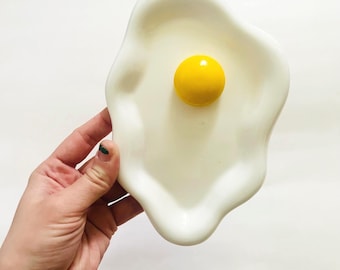 Puede incluir: Un plato de cerámica blanco con forma de huevo frito, con una yema amarilla brillante en el centro. El plato tiene un acabado liso y brillante y un diseño caprichoso. Es un artículo decorativo único.