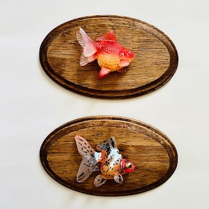 May include: Two framed, mounted, resin goldfish. The top goldfish is orange and red with a white tail fin. The bottom goldfish is orange, white, and black with a white tail fin. Both are mounted on oval, wooden plaques.
