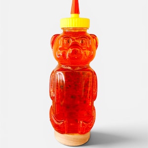 May include: A clear, bear-shaped plastic squeeze bottle filled with a vibrant orange liquid containing visible dark specks. The bottle features a bright yellow screw-on cap topped with a small, pointed red nozzle and rests on a light wooden base.