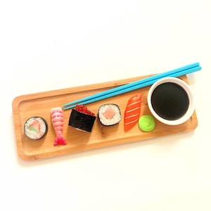 May include: A wooden sushi serving tray with various sushi pieces, a small white bowl of soy sauce, and blue chopsticks. The sushi includes a roll, shrimp, a dark roll with red roe, and a salmon piece. A small green ball of wasabi is also present.