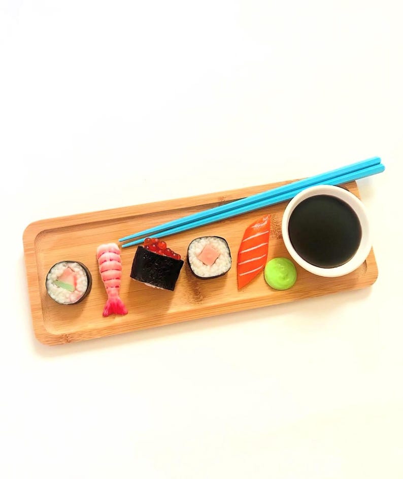 May include: A wooden sushi serving tray with various sushi pieces, a small white bowl of soy sauce, and blue chopsticks. The sushi includes a roll, shrimp, a dark roll with red roe, and a salmon piece. A small green ball of wasabi is also present.