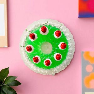 Peut inclure: Un dessert de gelée vert en forme d'anneau, garni de cerises rouges et de crème blanche. Le dessert est posé sur un fond rose, avec un bord décoratif blanc. L'image a une esthétique rétro et ludique.