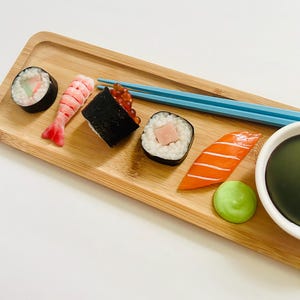 May include: A wooden serving tray with an assortment of sushi rolls, including a shrimp nigiri, a salmon nigiri, and a roll with red roe. Blue chopsticks rest on the tray, alongside a small bowl of dark sauce and a dollop of green wasabi.