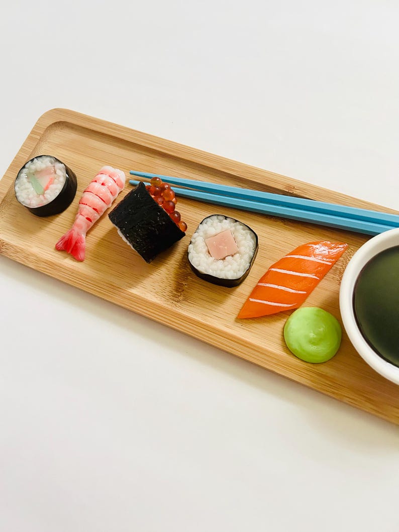 May include: A wooden serving tray with an assortment of sushi rolls, including a shrimp nigiri, a salmon nigiri, and a roll with red roe. Blue chopsticks rest on the tray, alongside a small bowl of dark sauce and a dollop of green wasabi.