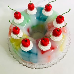 May include: A vibrant, ring-shaped dessert with a clear, jelly-like base. The dessert features a rainbow of colours and is decorated with swirls of white cream and bright red cherries with green stalks. The dessert is presented on a clear, scalloped-edge plate.