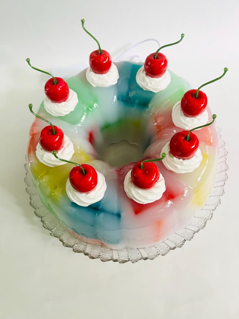 May include: A vibrant, ring-shaped dessert with a clear, jelly-like base. The dessert features a rainbow of colours and is decorated with swirls of white cream and bright red cherries with green stalks. The dessert is presented on a clear, scalloped-edge plate.
