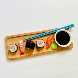 May include: A wooden sushi serving tray with various sushi rolls, nigiri, and a small bowl of soy sauce. Blue chopsticks rest on the tray. The sushi includes a shrimp, salmon, and other ingredients. A small green ball of wasabi is also present.