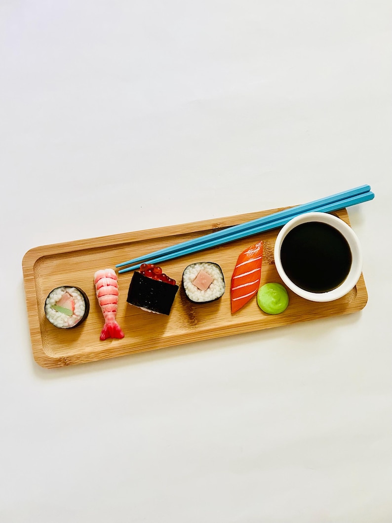 May include: A wooden sushi serving tray with various sushi rolls, nigiri, and a small bowl of soy sauce. Blue chopsticks rest on the tray. The sushi includes a shrimp, salmon, and other ingredients. A small green ball of wasabi is also present.
