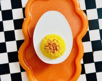 Puede incluir: Un plato de cerámica blanco en forma de huevo con un huevo relleno amarillo en su interior. El plato está colocado sobre una bandeja de color naranja brillante con un borde festoneado. La bandeja está sobre una superficie a cuadros en blanco y negro.
