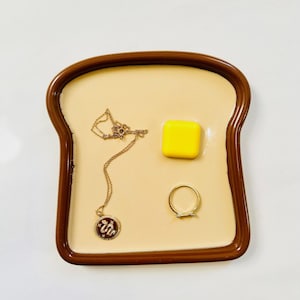 May include: A brown ceramic dish shaped like a slice of toast with a yellow butter pat, a gold necklace with a snake pendant, and a gold ring.