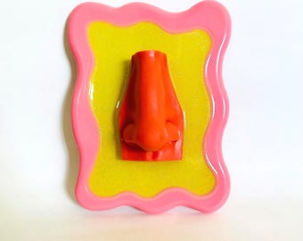 May include: A bright orange nose sculpture is centered on a yellow, glittery background, framed by a wavy pink border. The artwork is a unique, eye-catching piece, perfect for adding a pop of color and personality to any space.