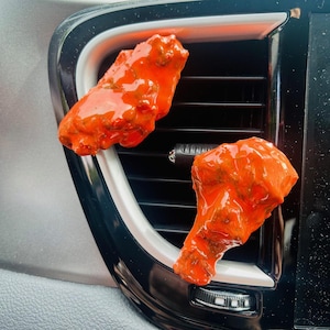 May include: Two bright orange, saucy chicken wings are placed on a car's air vent. The wings are glistening with sauce, and the air vent is black with a silver trim. The image is a humorous take on car accessories.