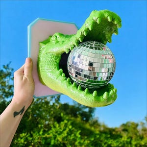 Pode incluir: Decora&ccedil;&atilde;o de parede em forma de jacar&eacute; verde segurando uma bola de discoteca. O jacar&eacute; &eacute; montado em uma base retangular rosa e azul. A bola de discoteca &eacute; redonda e coberta por pequenos quadrados reflexivos.