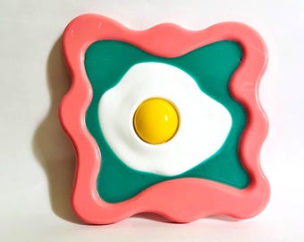 Puede incluir: Una pieza decorativa colorida con un diseño de huevo frito. El huevo tiene una base blanca con una yema amarilla, sobre un fondo verde azulado, y enmarcado por un borde ondulado de color coral.