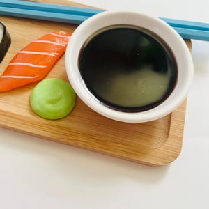 May include: A close-up of a sushi arrangement on a bamboo tray. The arrangement includes a salmon nigiri, a dollop of wasabi, and a small white bowl filled with dark soy sauce. Blue chopsticks are placed on the tray.