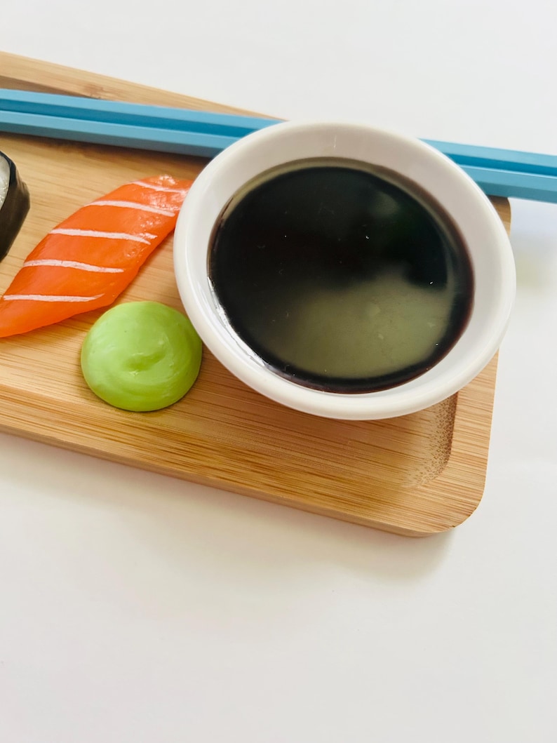 May include: A close-up of a sushi arrangement on a bamboo tray. The arrangement includes a salmon nigiri, a dollop of wasabi, and a small white bowl filled with dark soy sauce. Blue chopsticks are placed on the tray.