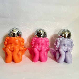 May include: Three cherub figurines in orange, pink, and lavender, each topped with a silver disco ball. The cherubs rest their chins on their hands, with small wings visible. The background is a plain white.