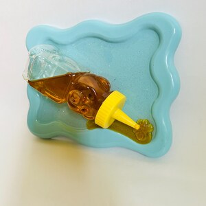 May include: A clear glass bottle with a yellow cap filled with honey is tipped over on a light blue tray with a wavy edge. The honey has spilled out of the bottle and onto the tray.