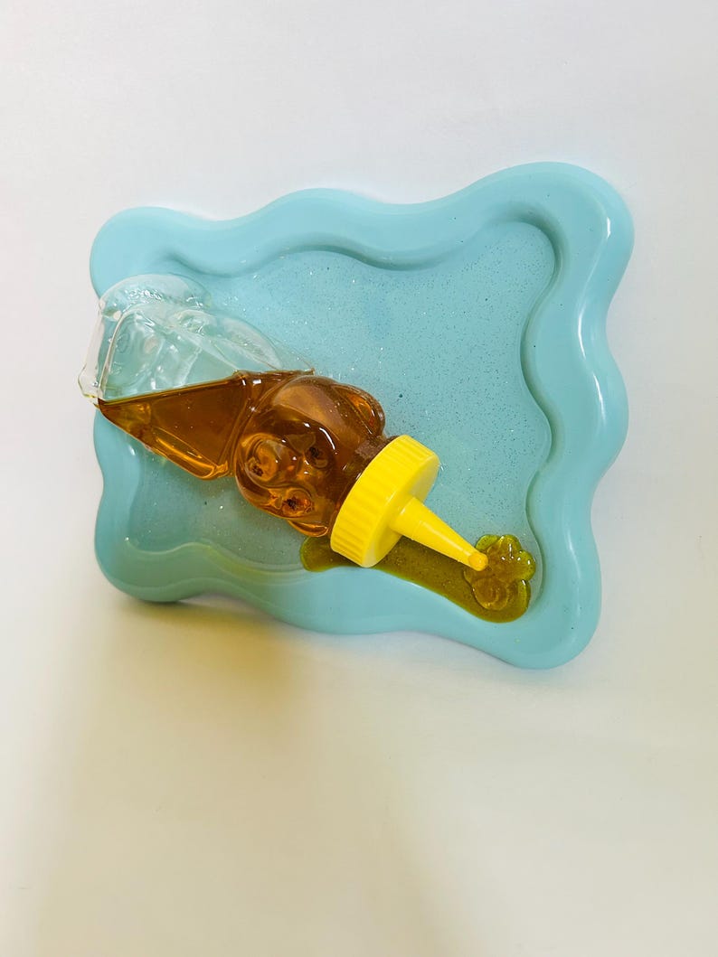 May include: A clear glass bottle with a yellow cap filled with honey is tipped over on a light blue tray with a wavy edge. The honey has spilled out of the bottle and onto the tray.